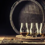 Glencairn whiskey tasting cups on a wooden serving, with a whisky barrel in the dark background